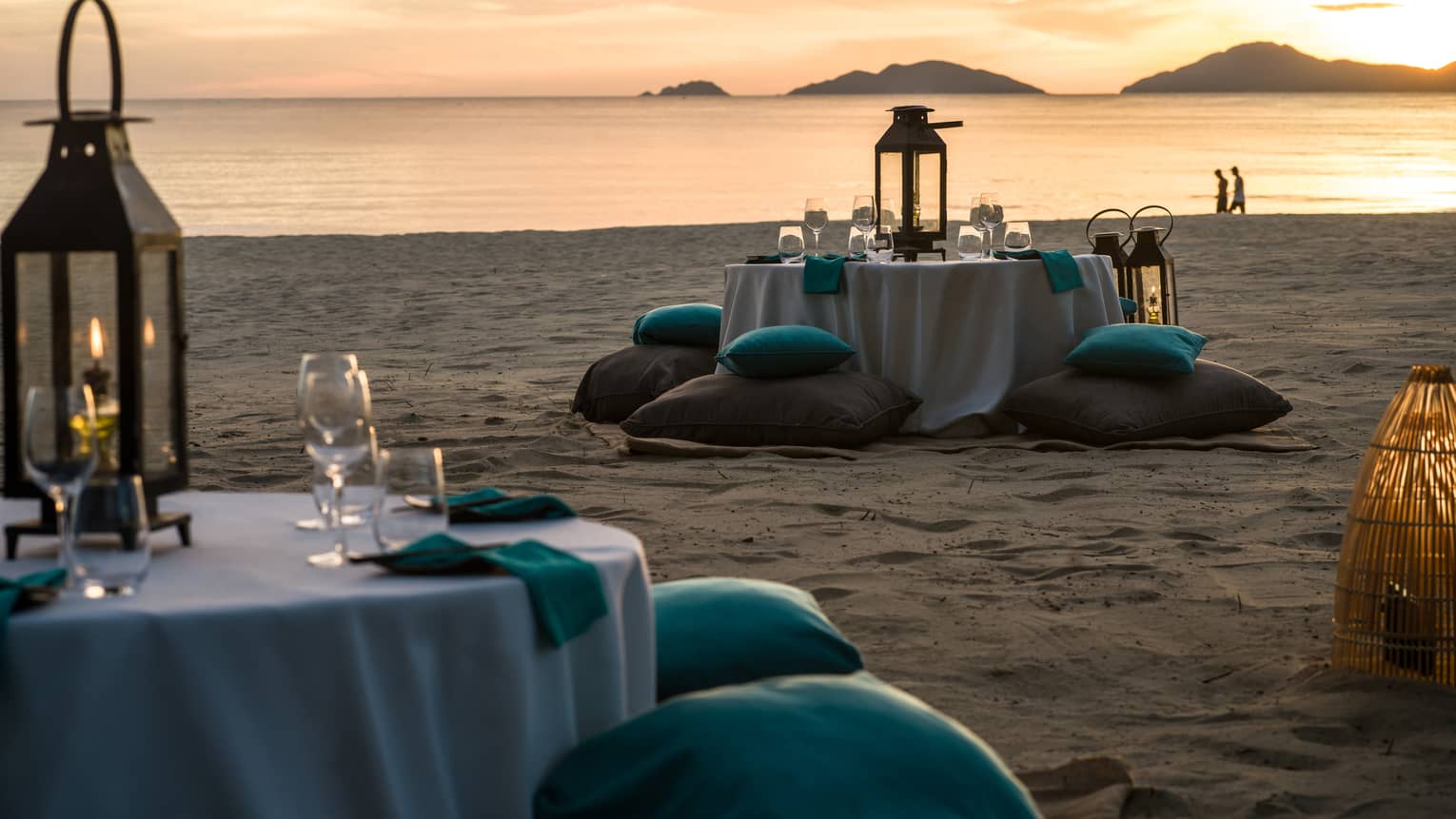 Two tables with white cloths, glasses, lanterns and turquoise cushions on sandy beach at dusk 