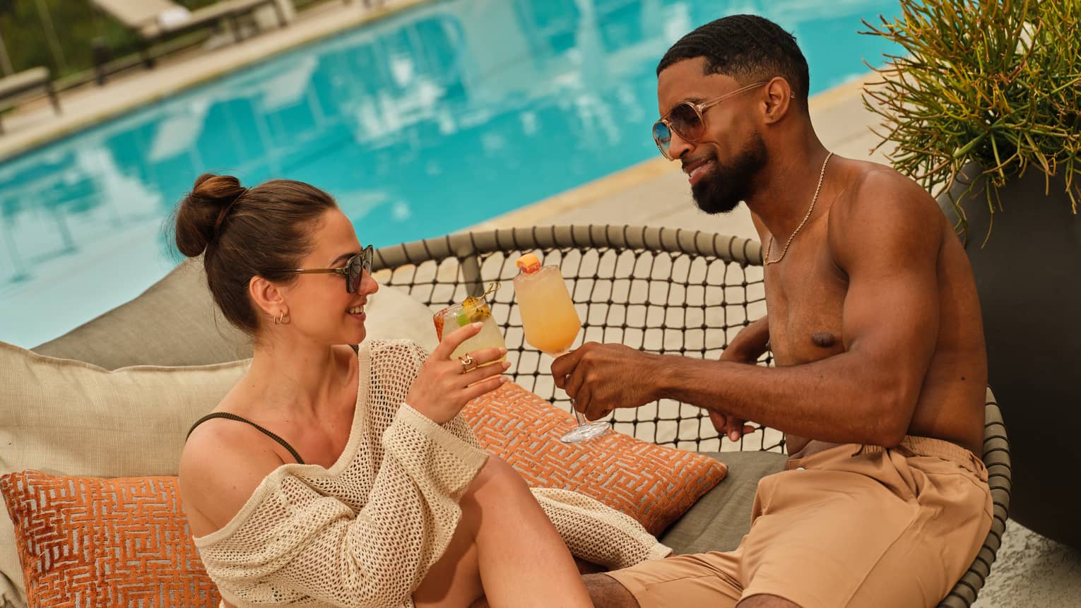 A couple toasts cocktails on a poolside lounge chair