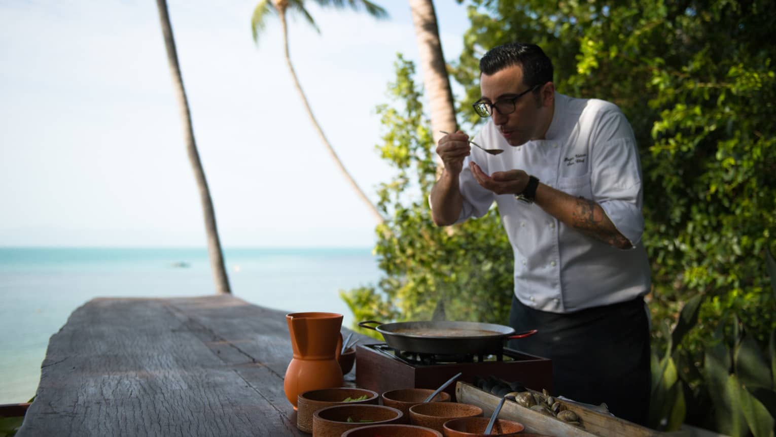 Chef in white coat blows on spoon in front of hot pot in outdoor kitchen