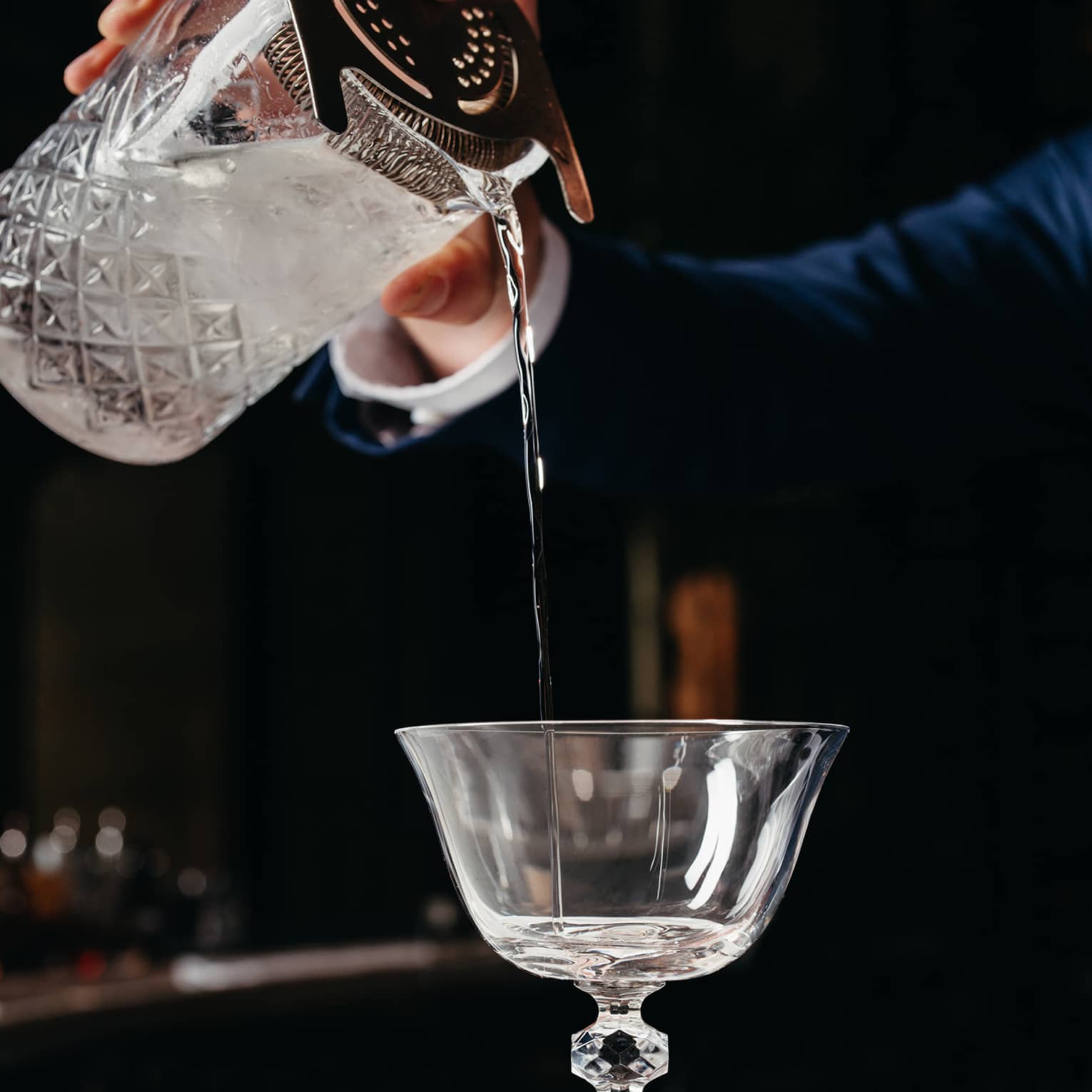 Hands pouring liquid from an ornate glass container into a cocktail glass