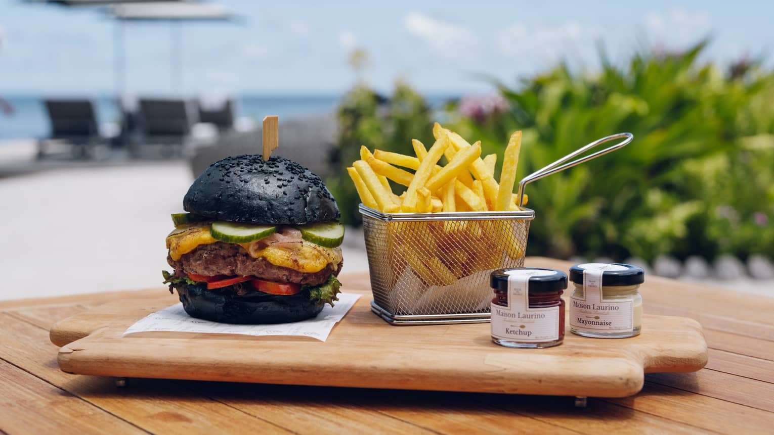 Burger with black bun and fries in metal basket on wooden serving board by the ocean