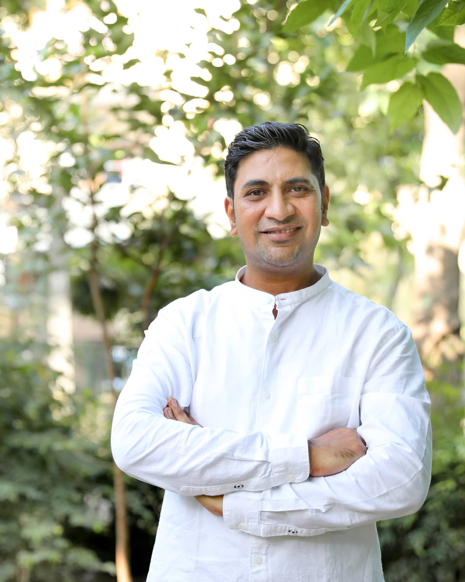 Smiling person in long-sleeved white linen shirt stands outdoors with arms crossed