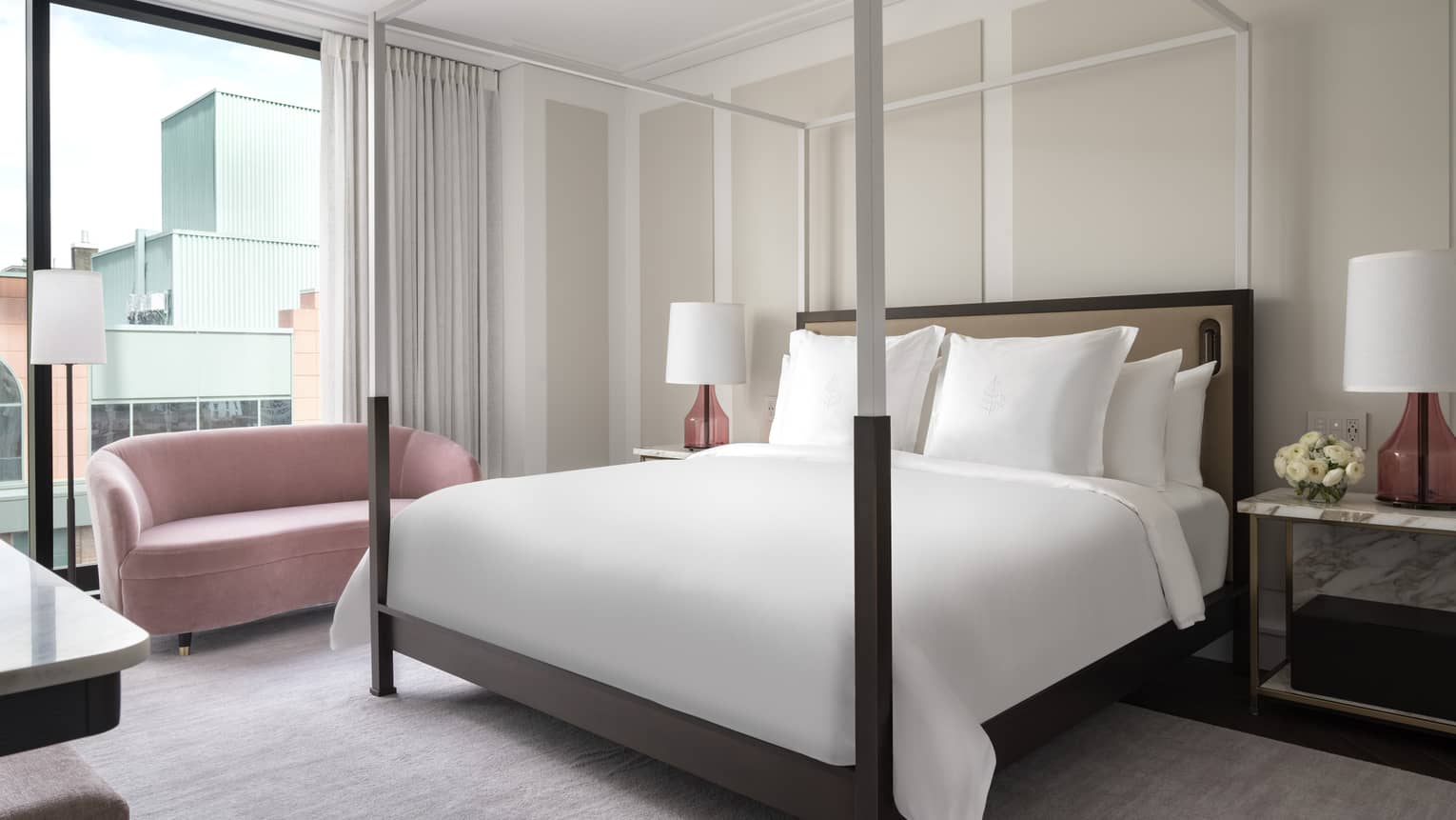A modern hotel bedroom houses a four-poster bed, white linens and pink accent lamps. A pink loveseat sits near large windows, offering views of adjacent buildings.