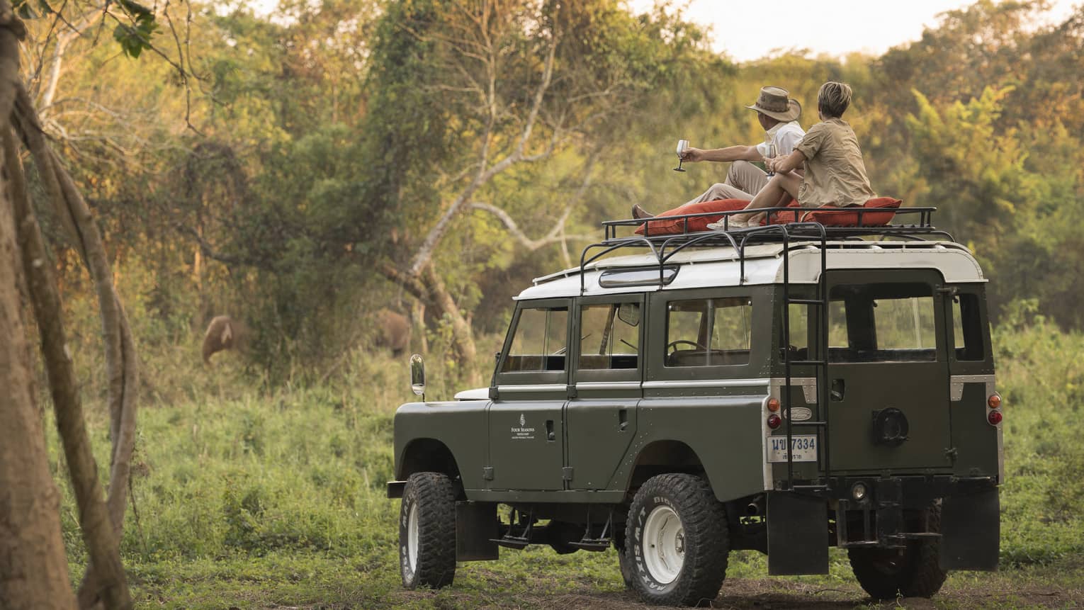 A couple sits on the roof of a Land Rover in the Bush, drinking a cocktail