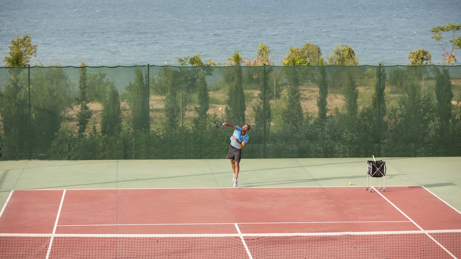 Man in blue shirt and black shorts serving a tennis ball on a seafront tennis court