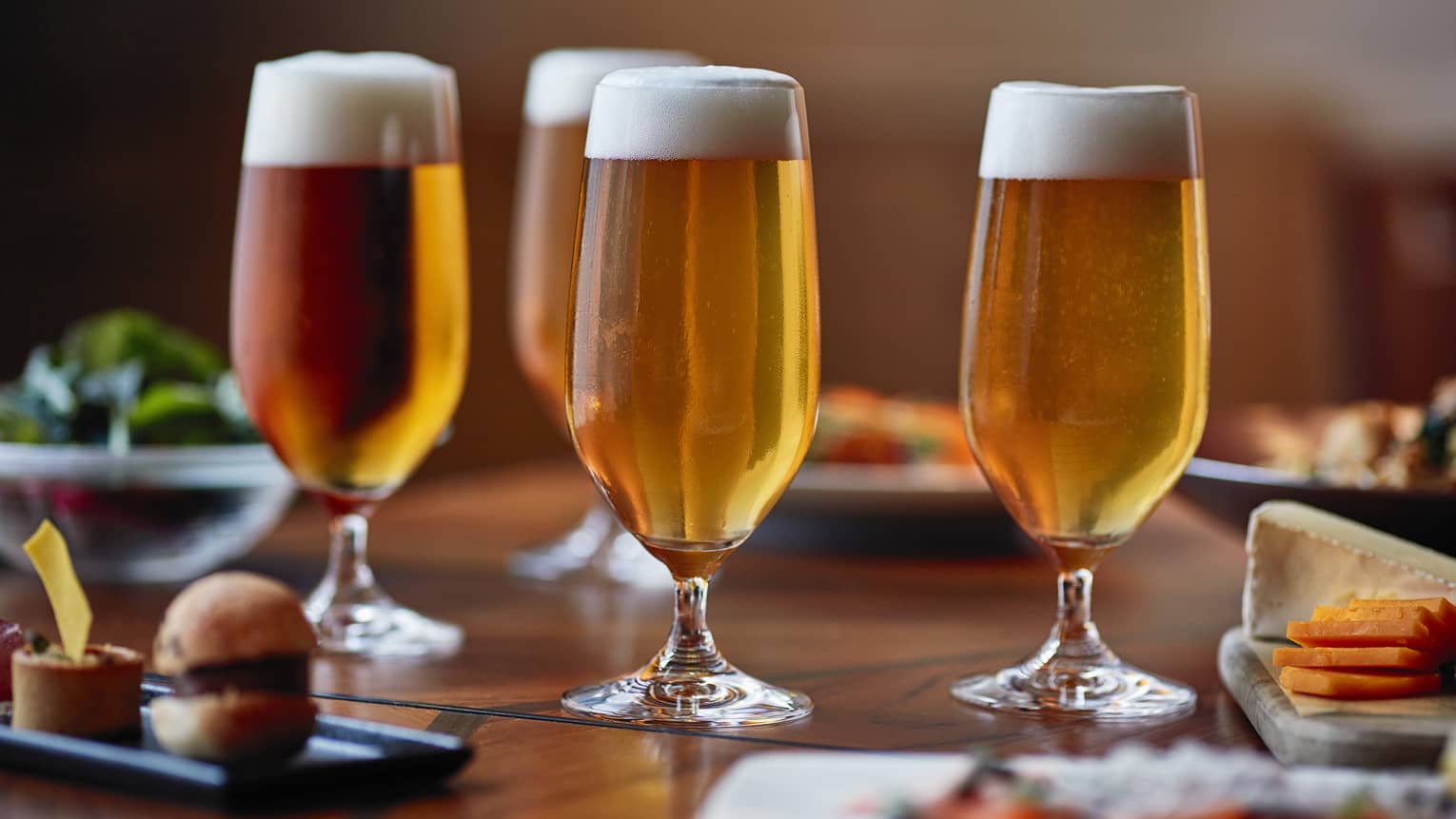 Four tall beer glasses filled with beer on a table with cheese, crackers and appetizers