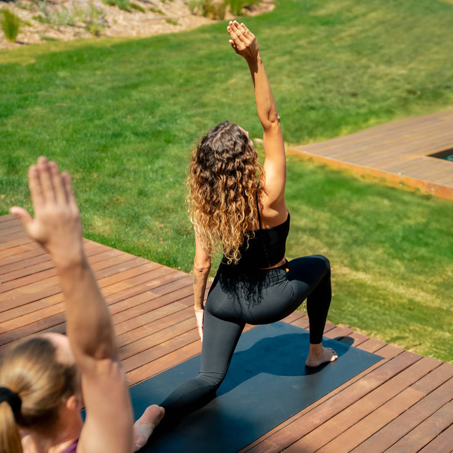 Two people in yoga pose practise on outdoor wooden deck overlooking lush green grass