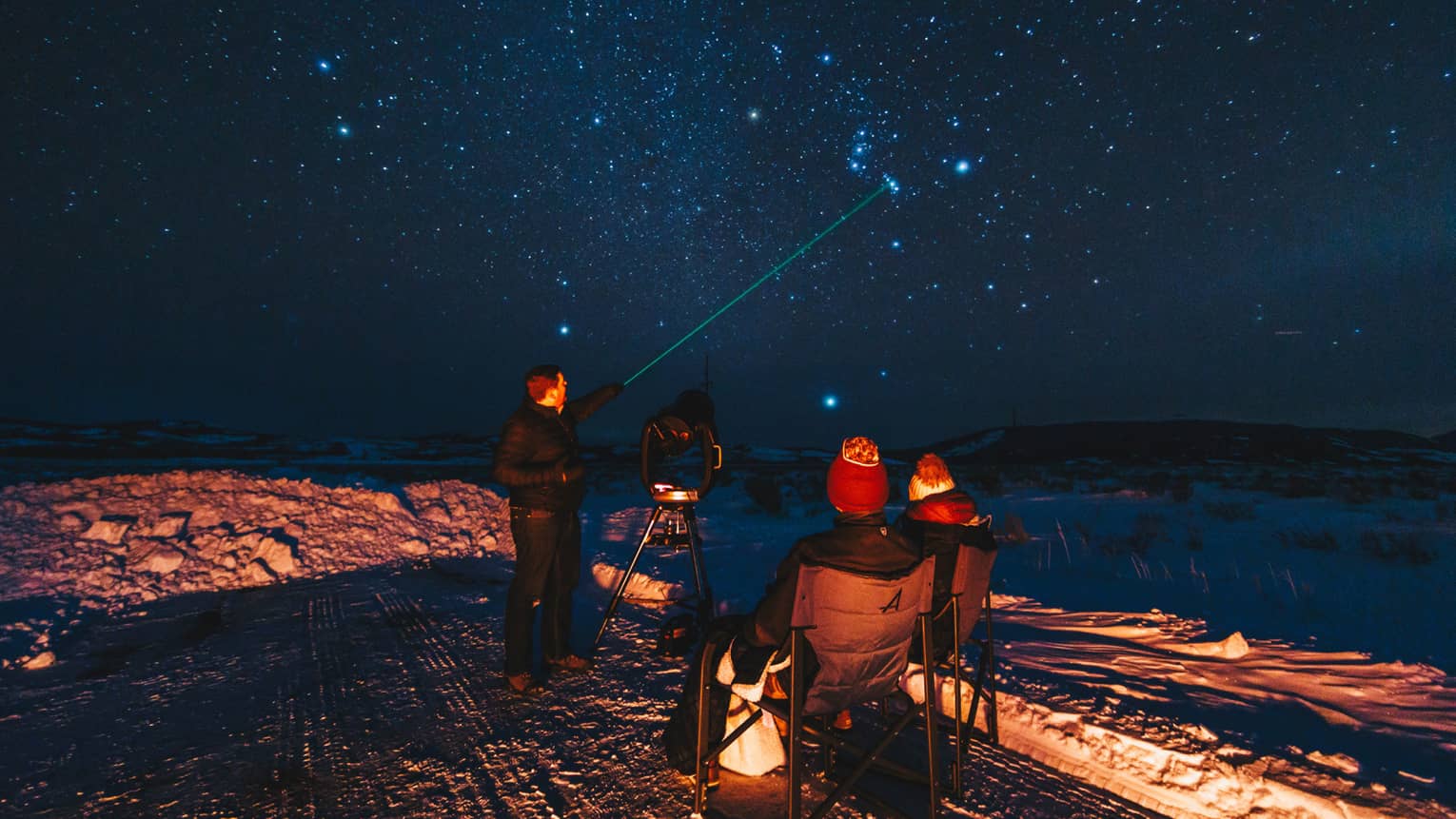 Group of people stargazing on a snowy night, with one person pointing a green laser at the sky, surrounded by chairs and a telescope 