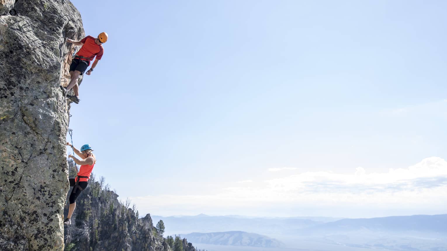 Man and woman climb steep cliff face with view of mountains in far distance 