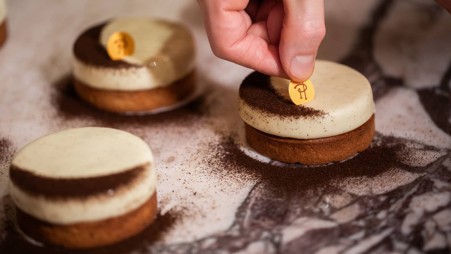 Three vanilla tarts topped with a crescent-shaped dusting of chocolate sit on a white table as a hand places a logo on a tart
