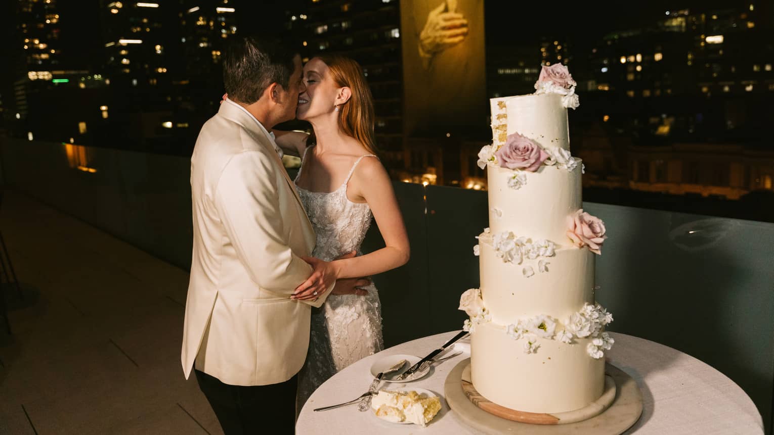 Woman wearing white sleeveless wedding dress and man wearing white tuxedo coat and black pants kiss on a rooftop terrace next to a wedding cake