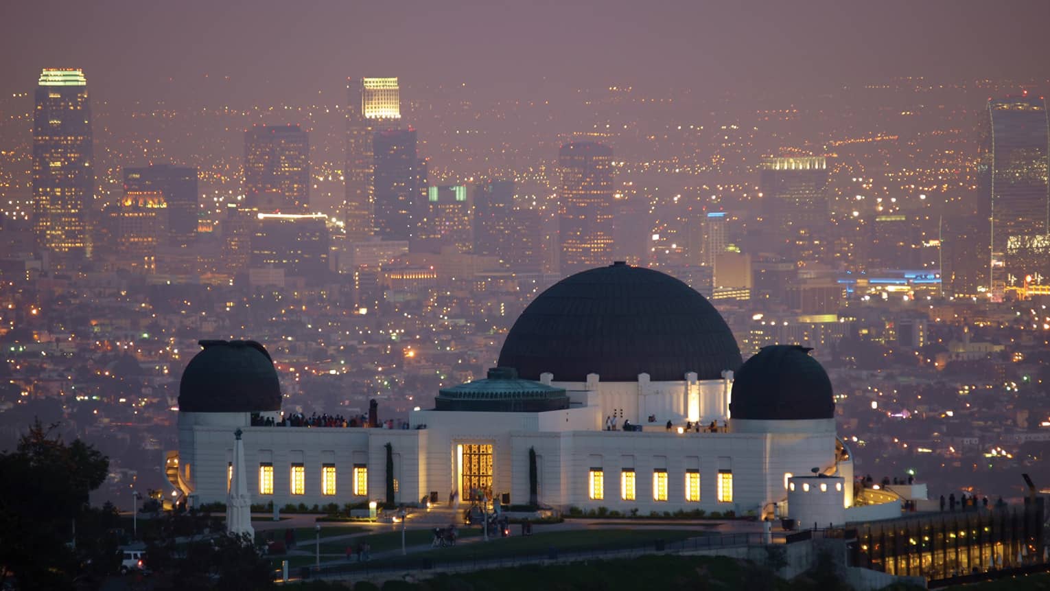 A large black and white observatory with three dome roofs, the city is behind it with many lights.