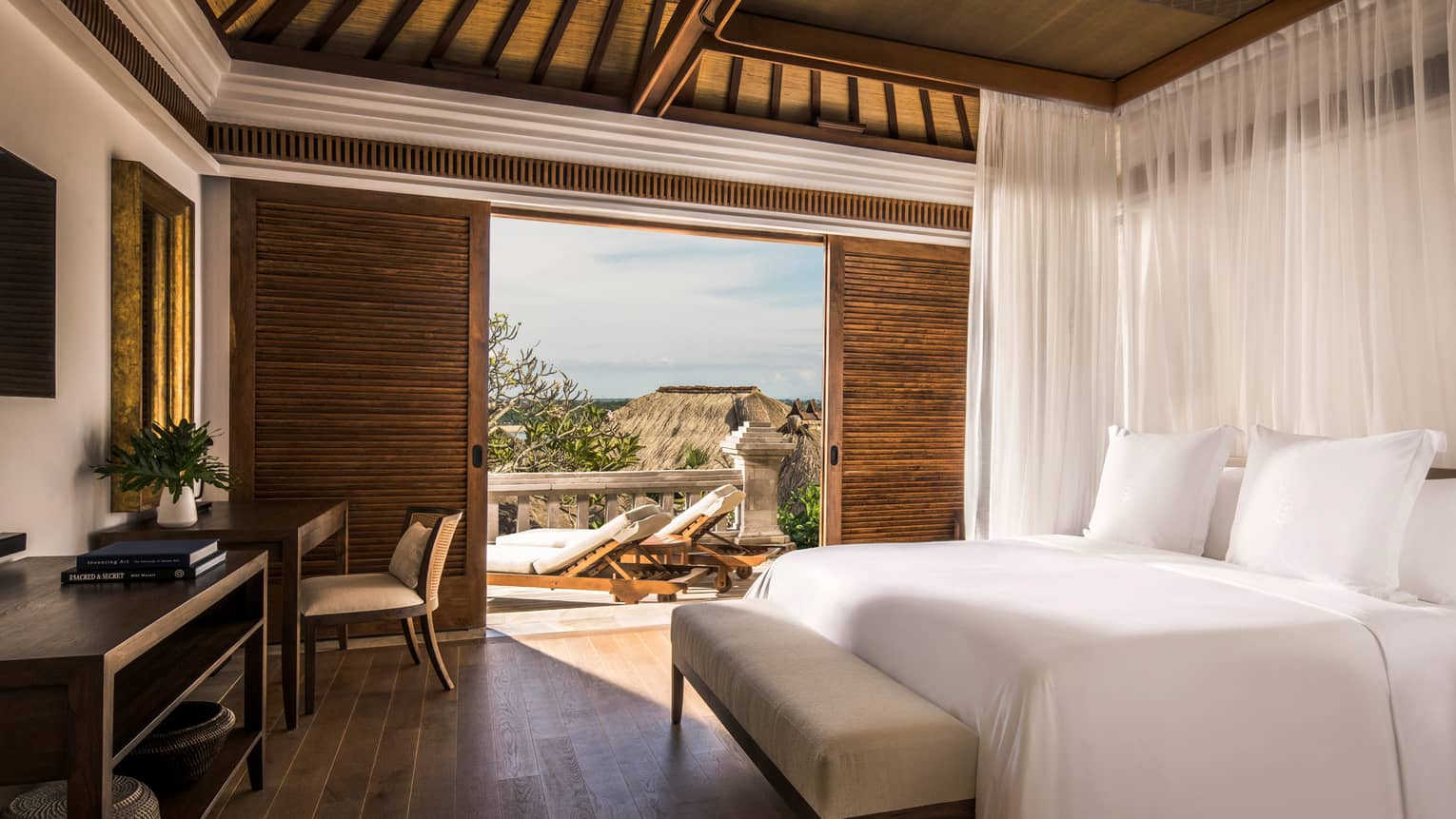 Garden villa bedroom bed, small bench, wood desk beside sliding wood shutters to sunny patio with garden view