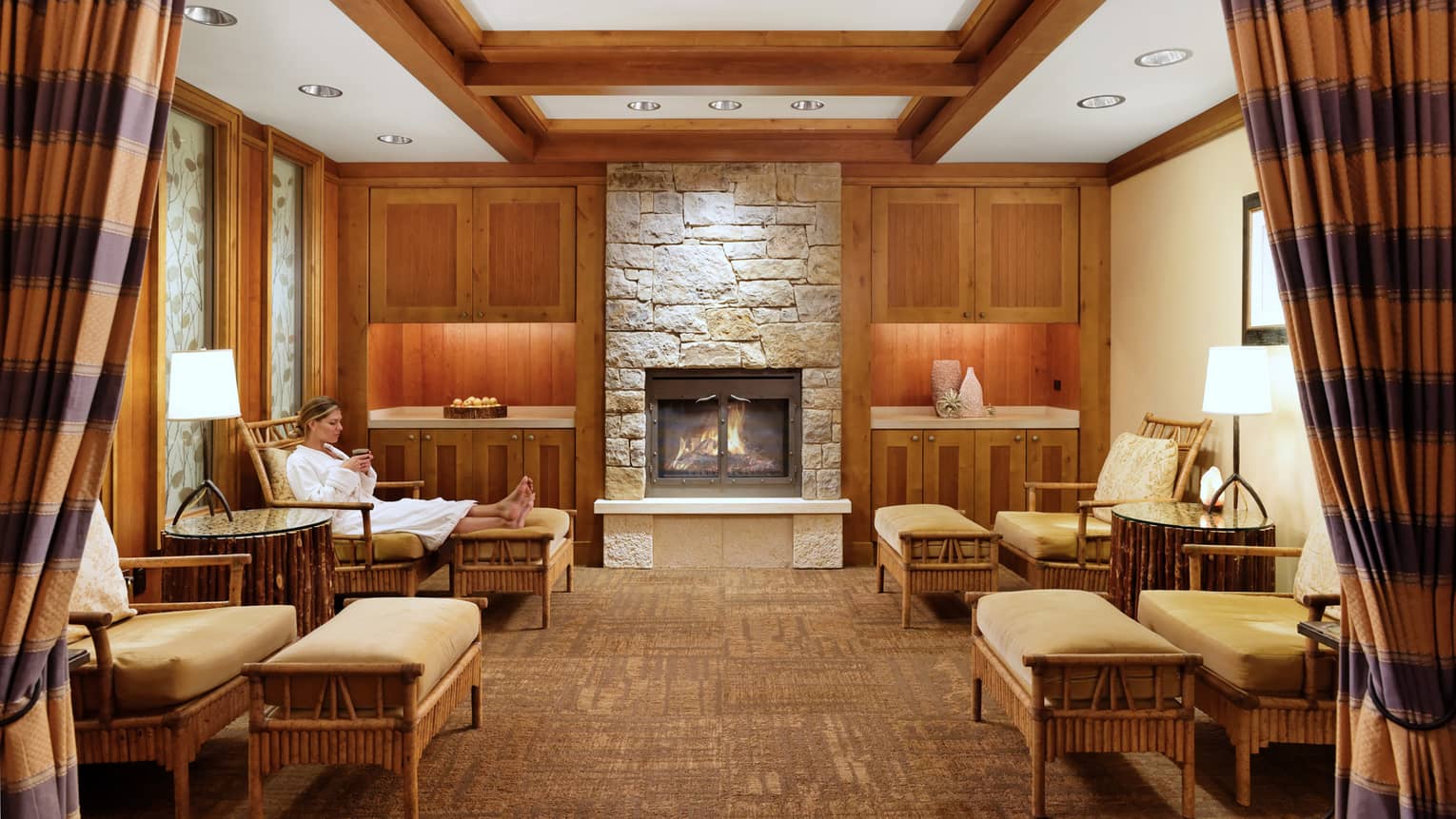 A woman relaxes on a cushioned chair by a lit fireplace