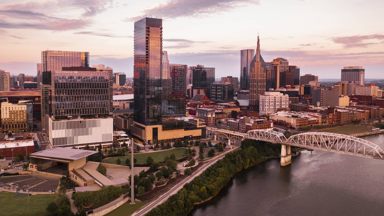 Aerial view of downtown Nashville, Tennessee at sunset, featuring modern skyscrapers, a river, a bridge, and a park with greenery in the foreground.