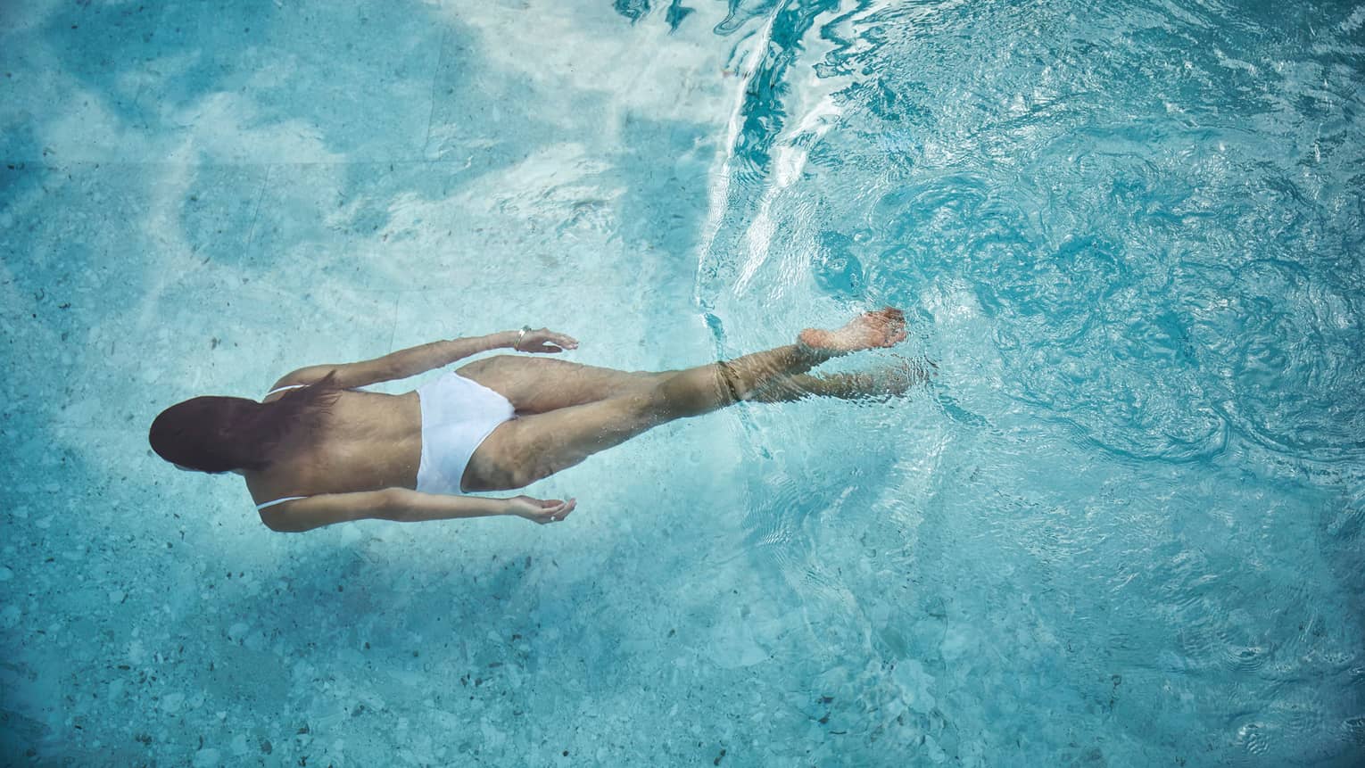 Woman in white swimsuit swimming underwater in blue pool