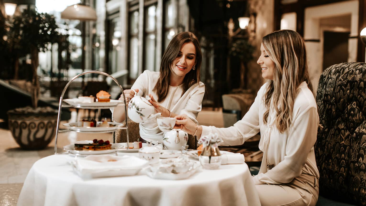 Two women in neutral outfits seated, smiling over afternoon tea in hotel lobby bar