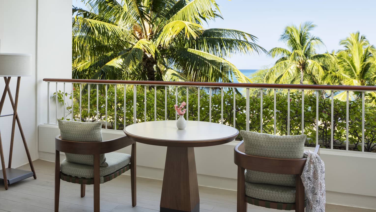 A private hotel balcony with two wooden chairs and a round table, facing palm trees and ocean