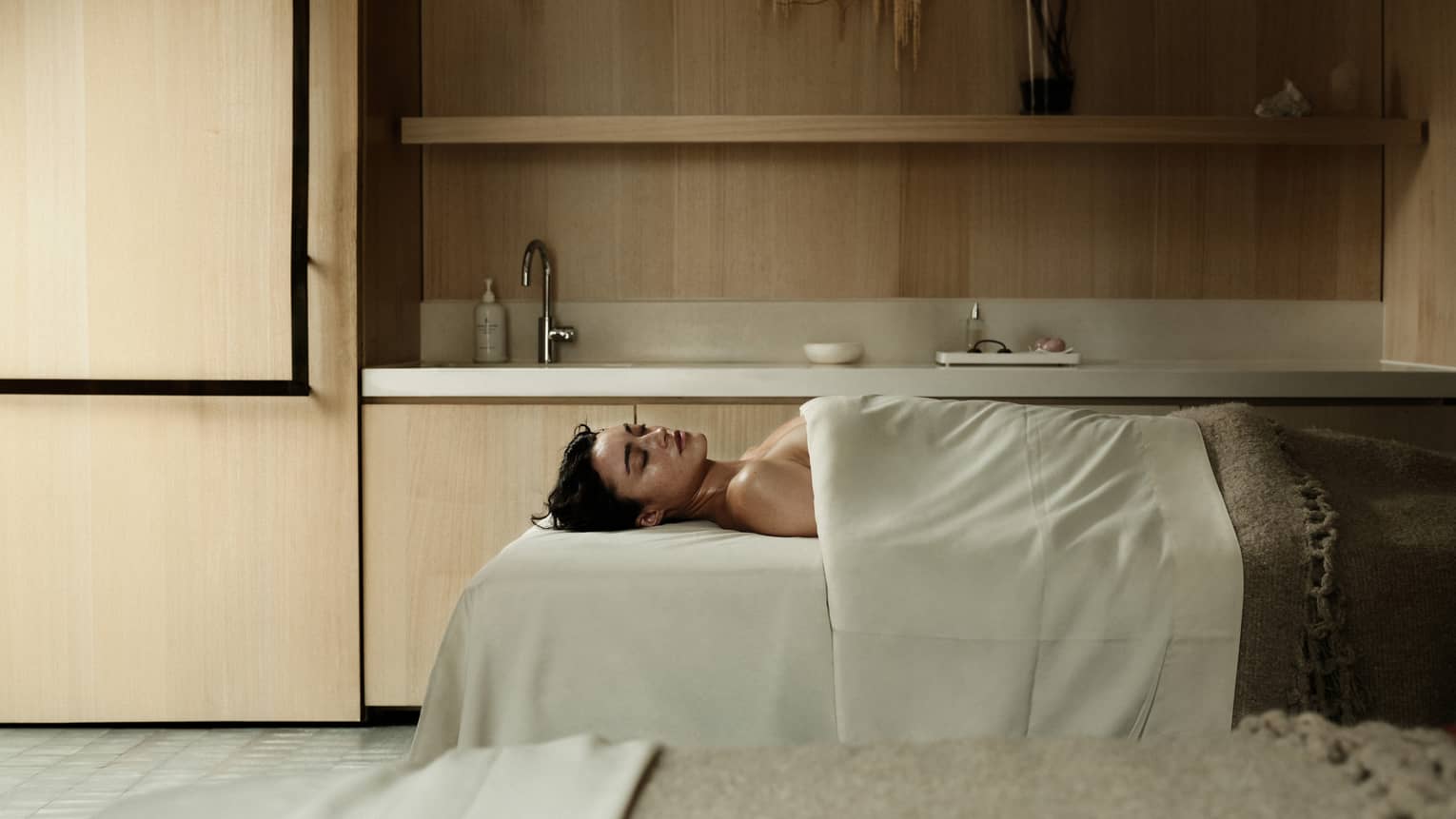 A person lies on supine on a spa treatment table in a serene, spacious treatment room and is draped with with a sheet and blanket.