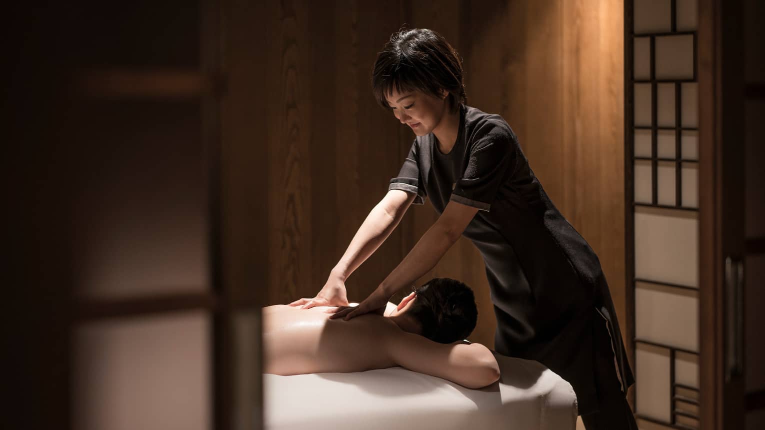 Man lies face-down on massage table as masseuse in black uniform rubs his shoulders