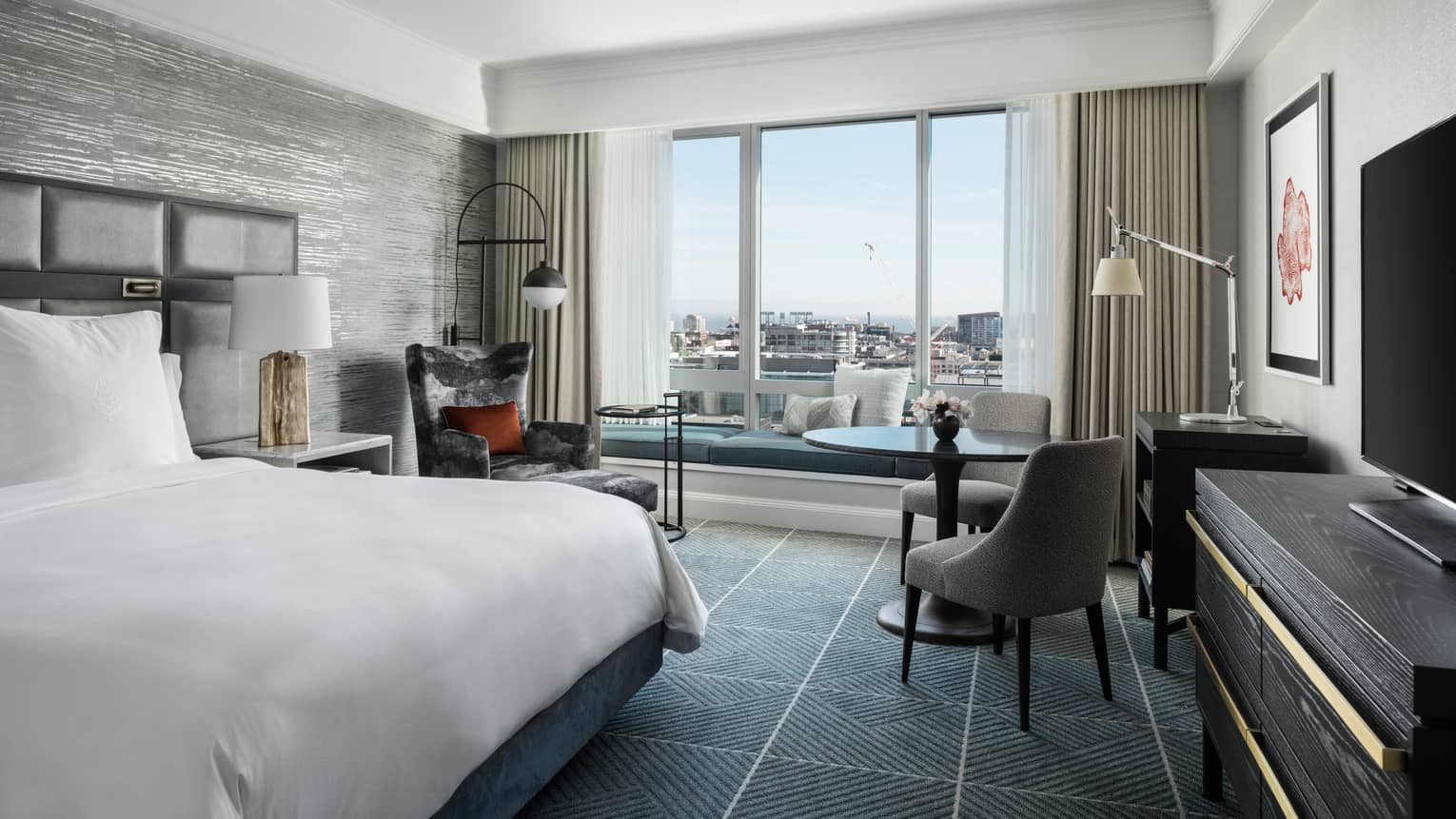Modern hotel room with king bed, table and two upholstered grey chairs, windows overlooking San Francisco