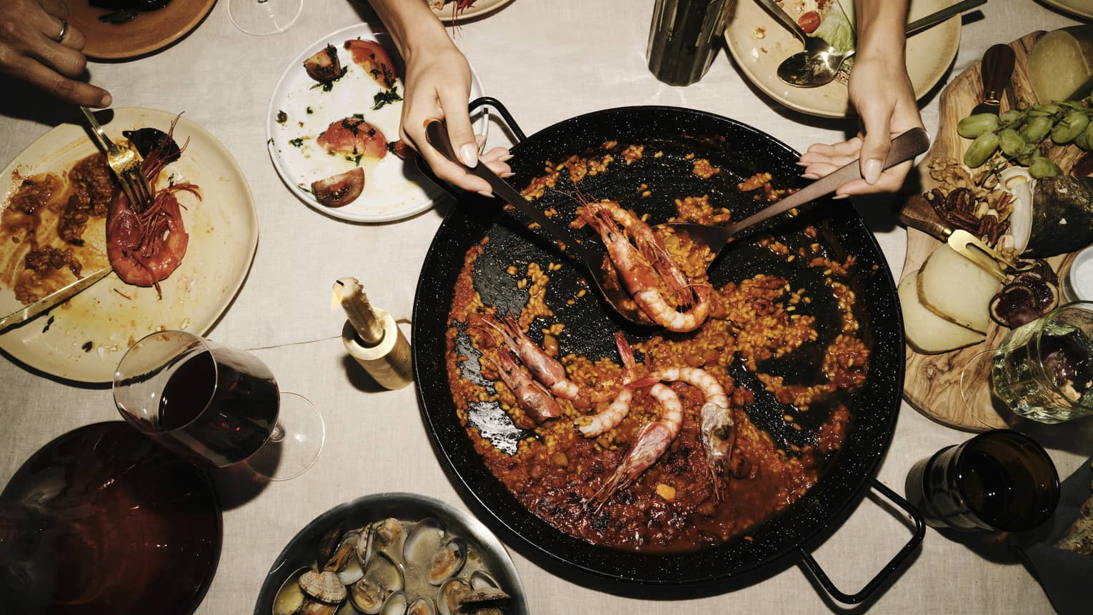 Two hands use large spoons to lift prawns from a saucy paella dish set amid plates of seafood, cheese and fruit.