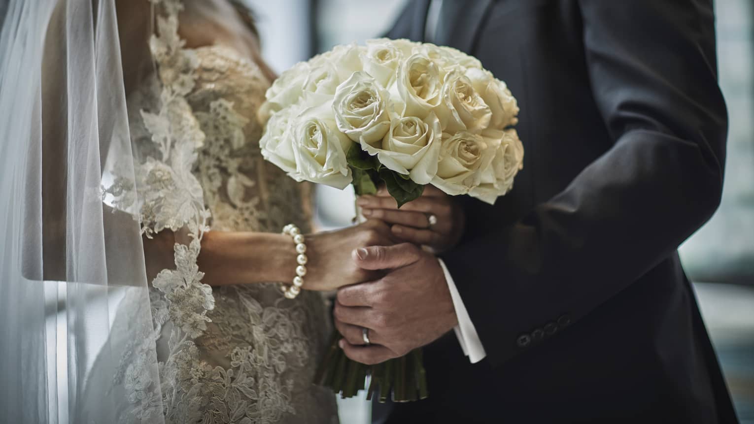 A close up of a bride in a lace gown and a groom in a gray suit hold the bride's bouquet of white roses together.