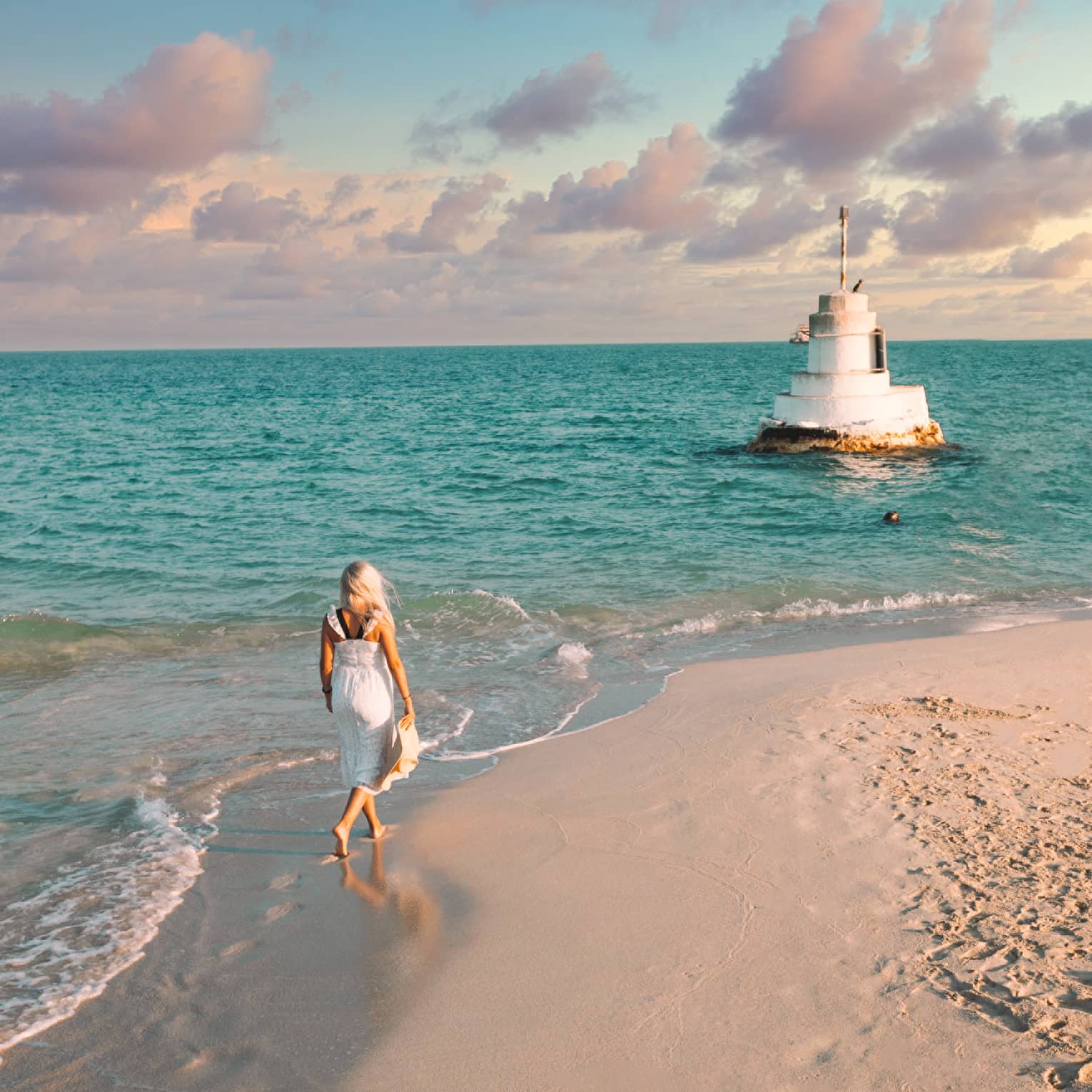 A woman walks along the shoreline towards a stone structure in the sea.