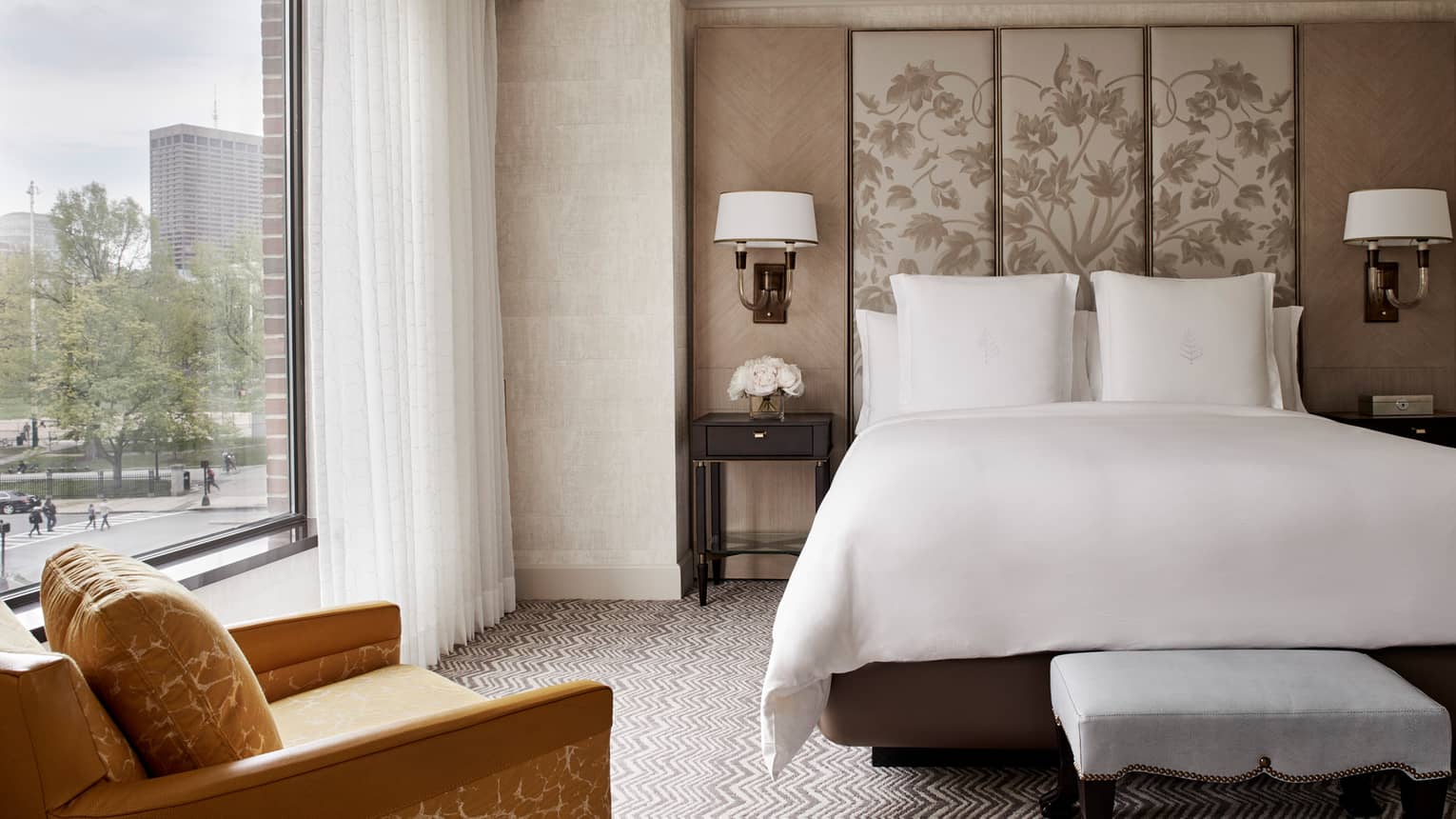 Front view of Executive Suite bed with white linens, beige fabric floral headboard, large window with white curtains, yellow armchair