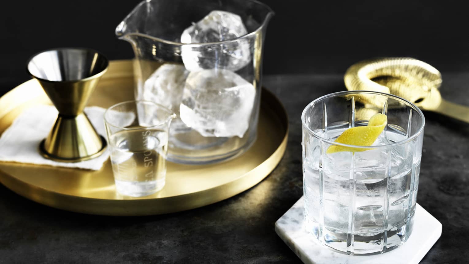 Brass platter, glass jug with ice cubes by rock glass with cocktail, lemon peel garnish