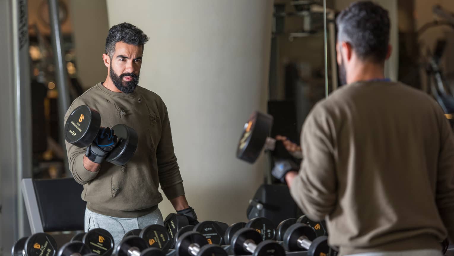 Man with beard wearing casual sweatshirt and sweatpants stands looks in gym mirror, lifts large hand weights