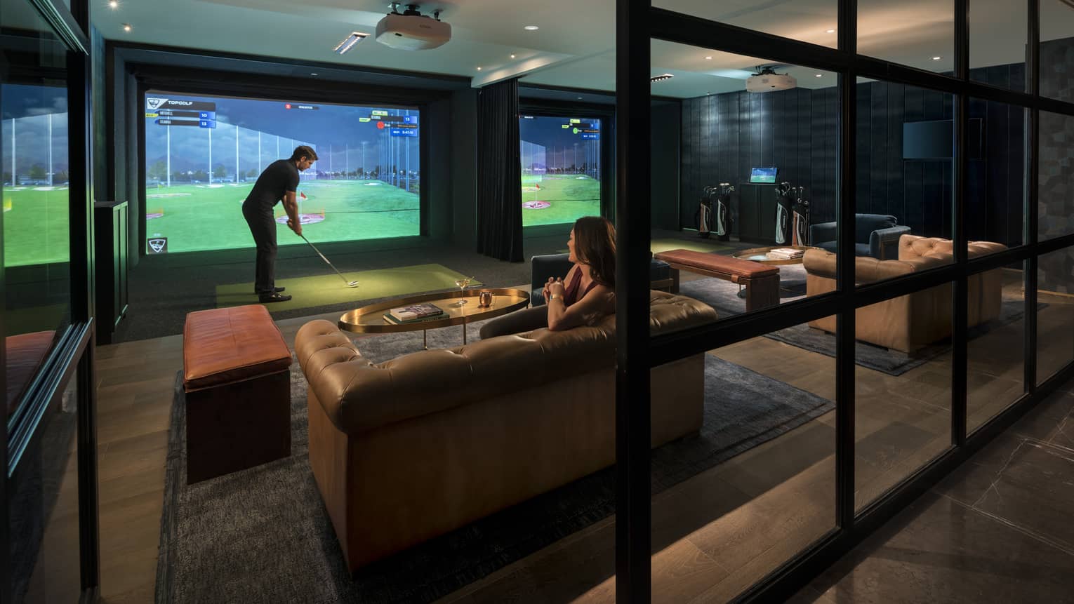 A man uses an indoor golf simulator while a woman watches from a nearby couch