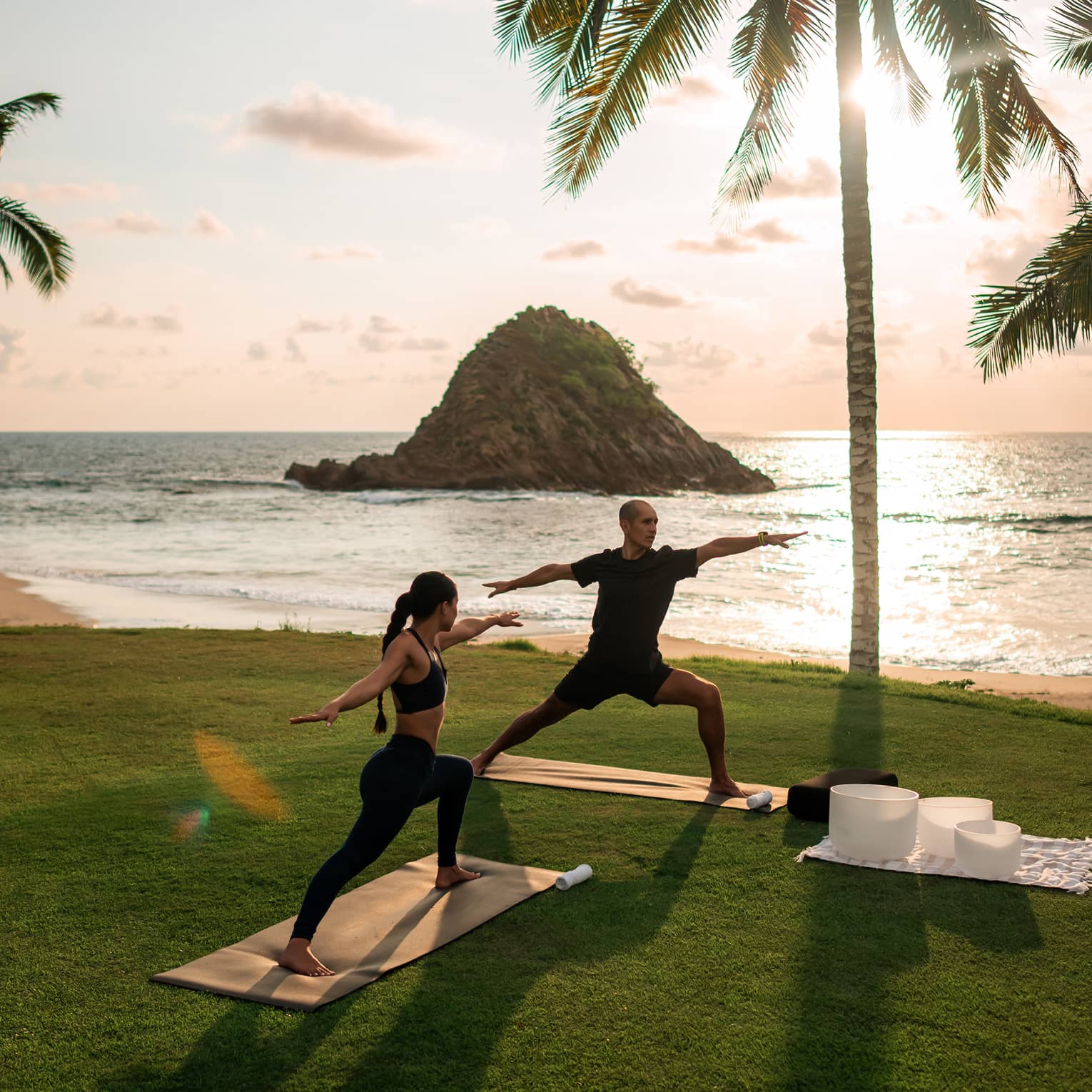 Two people in yoga poses on yoga mats on a lush grassy area next to the beach. A palm tree, the beach, the ocean and a bright but partially cloudy sky are in the background.