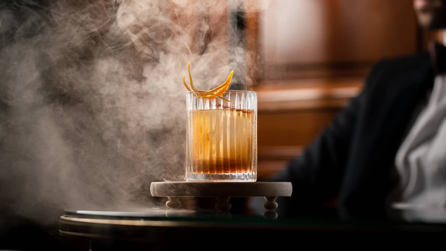 Smoke billows in the air as a glass dome is being lifted off of a whiskey-based cocktail in a rocks glass garnished with orange peel