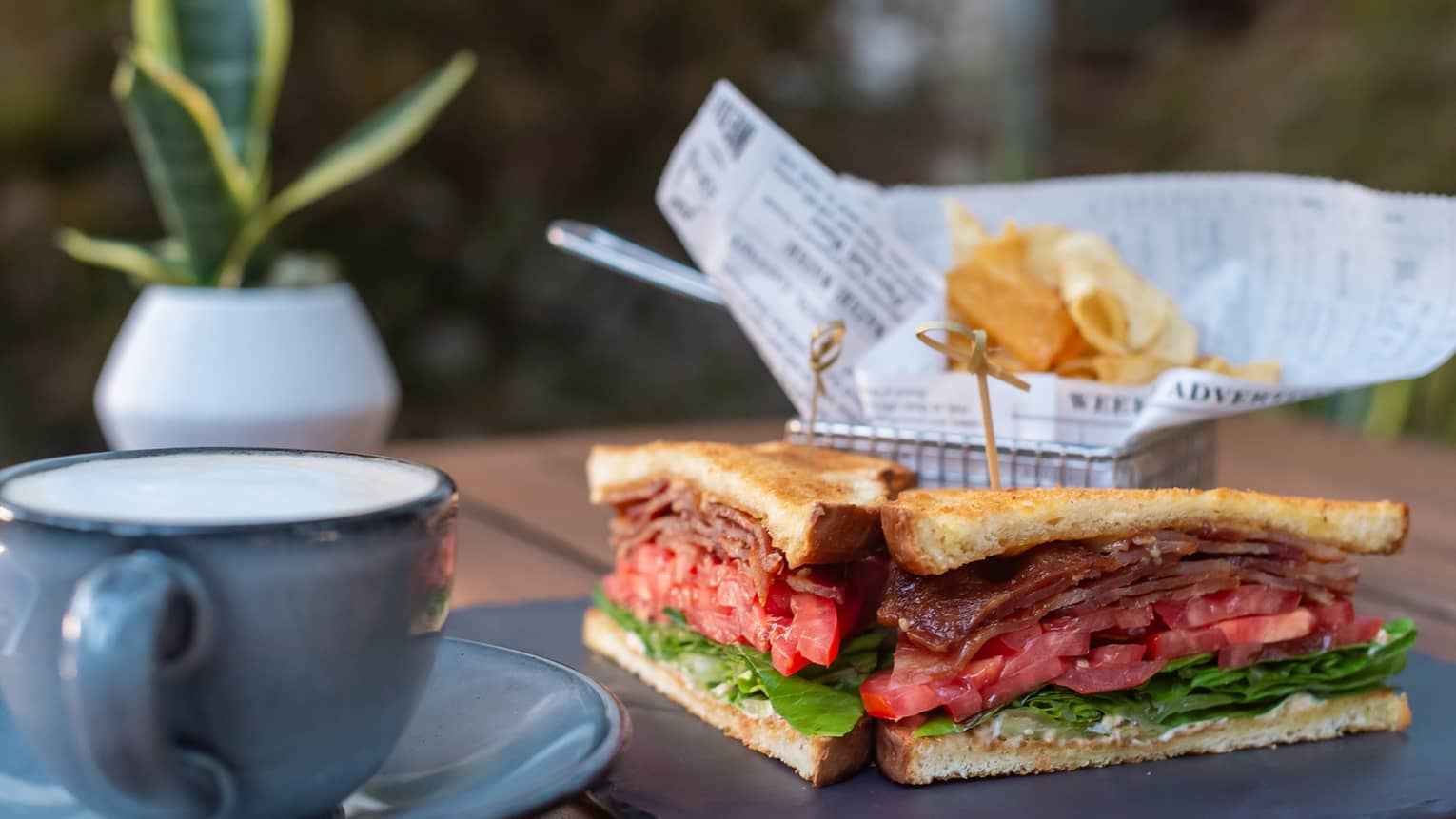 BLT sandwich cut in half, blue coffee cup and saucer, basket of chips