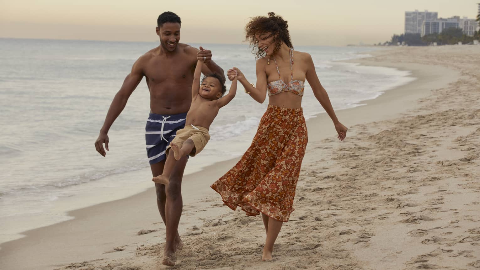 Smiling man and woman in beach attire swing young child by their arms as they walk on a Fort Lauderdale beach at sunset