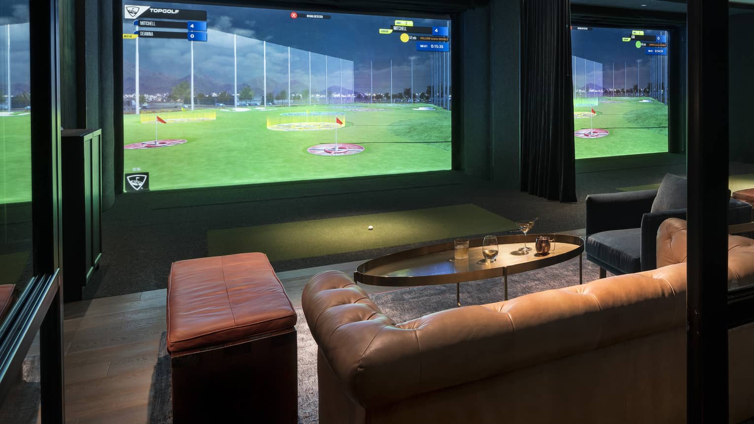 A golf simulator in a dimly lit room with a large screen, leather sofa, bench, and round coffee table.