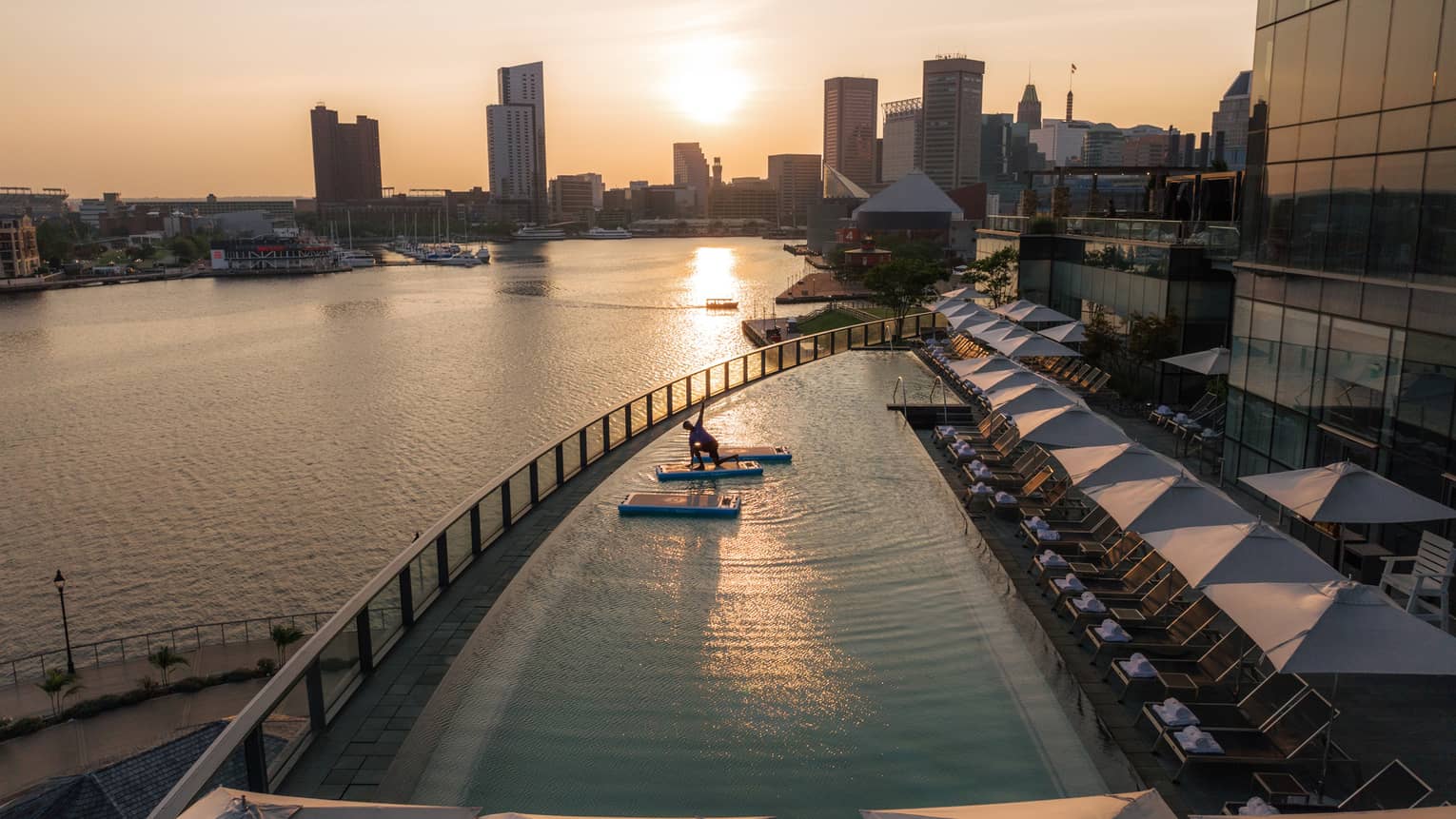 A guest practises yoga on a mat floating in a pool overlooking a river. In the background, the sun sets behind skyscrapers.