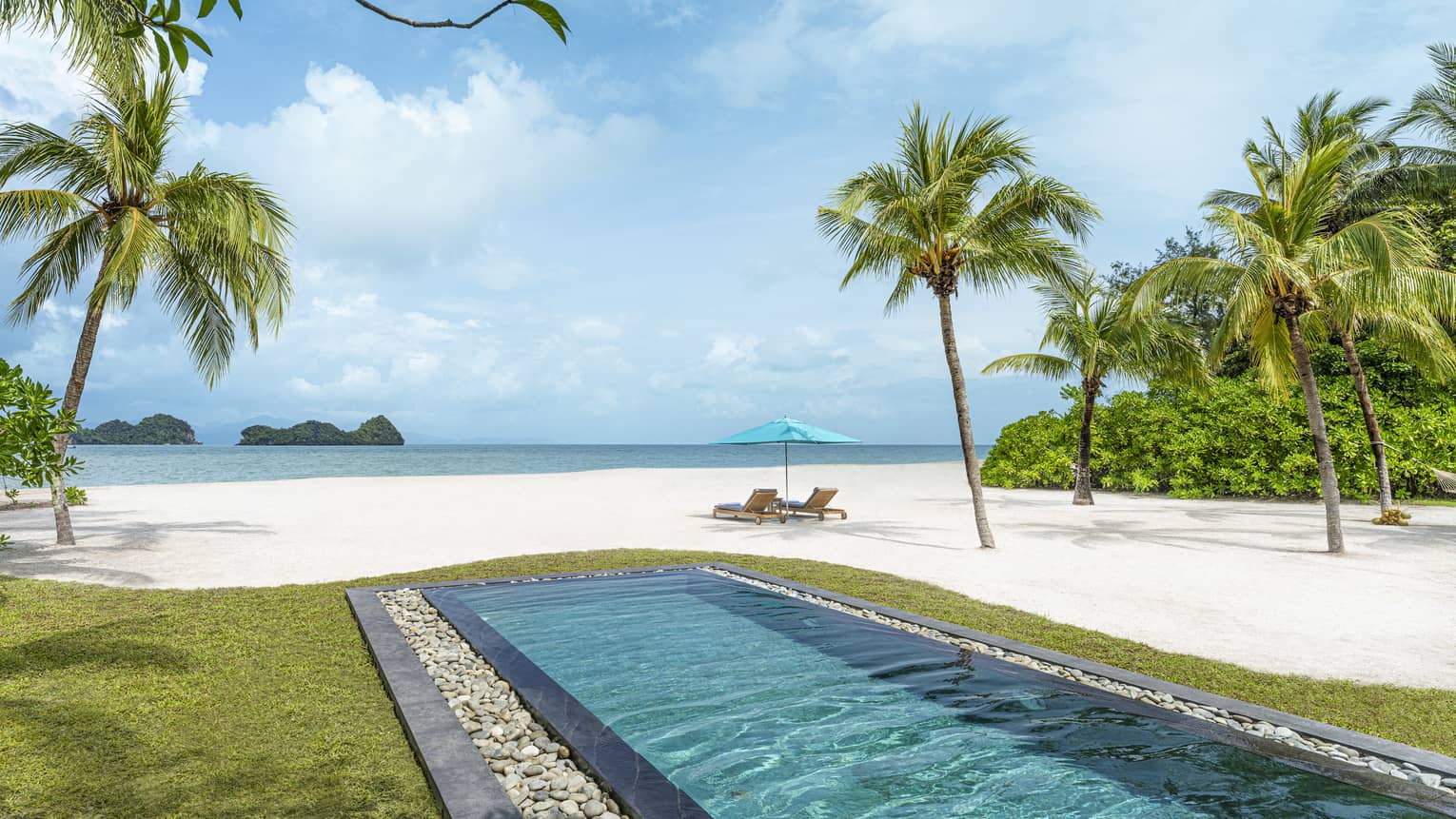 Rectangular in-ground pool jutting out to white-sand beach dotted with palm trees with two lounge chairs and umbrella along blue waters