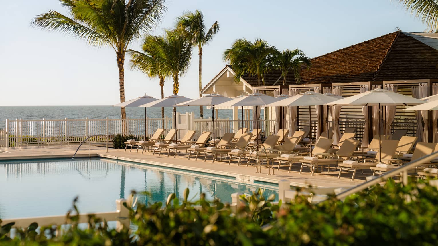 Outdoor pool lined with white lounge chairs an umbrellas with the beach in the background