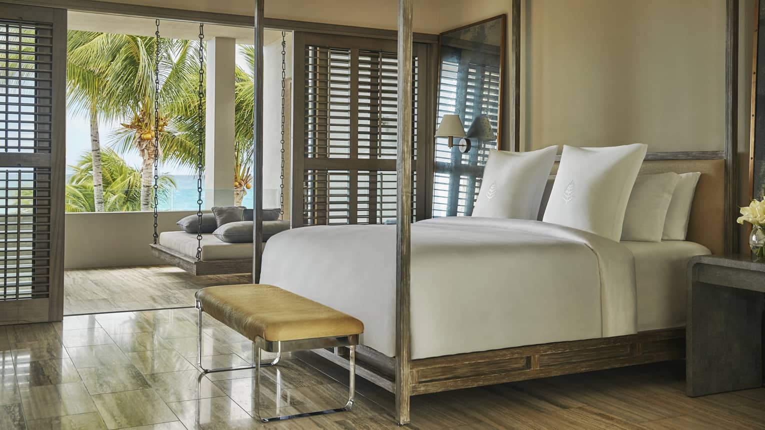 Bed with white linens, rustic wood canopy, small leather bench, open patio door shutters to hanging chair