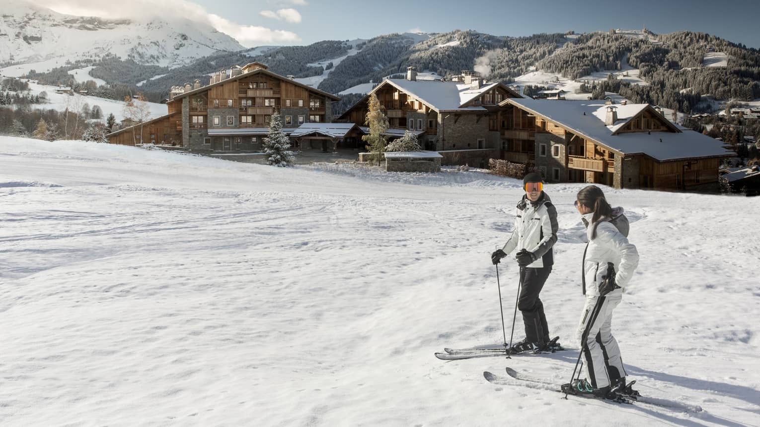 Man, woman on skis at top of snowy hill, Four Seasons Hotel Megeve in background