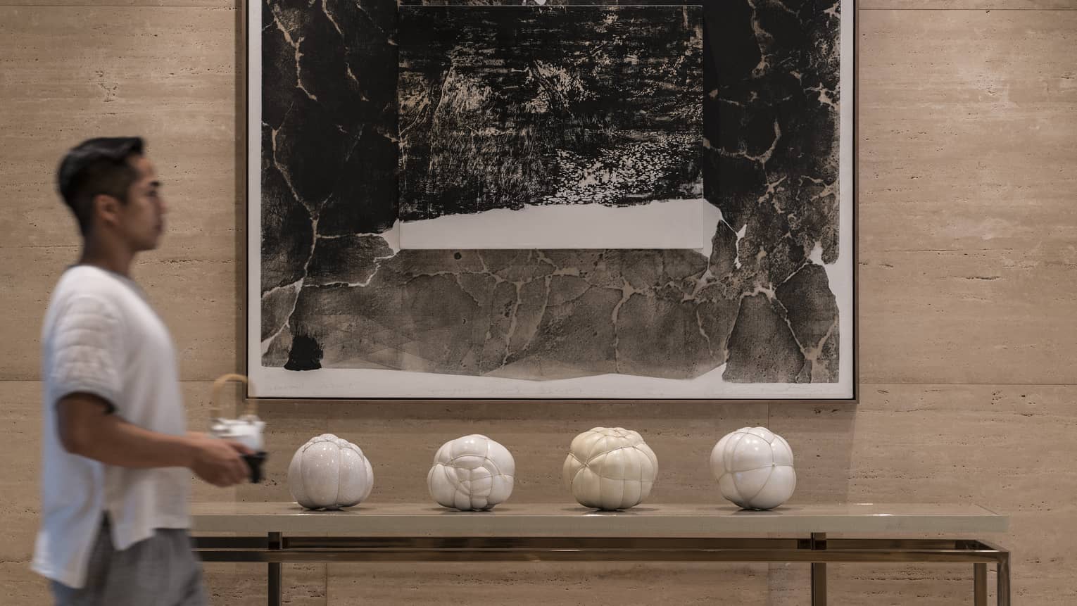 A blurred adult carrying a white teapot walks past a large abstract black-and-white painting hanging on a grey stone wall.