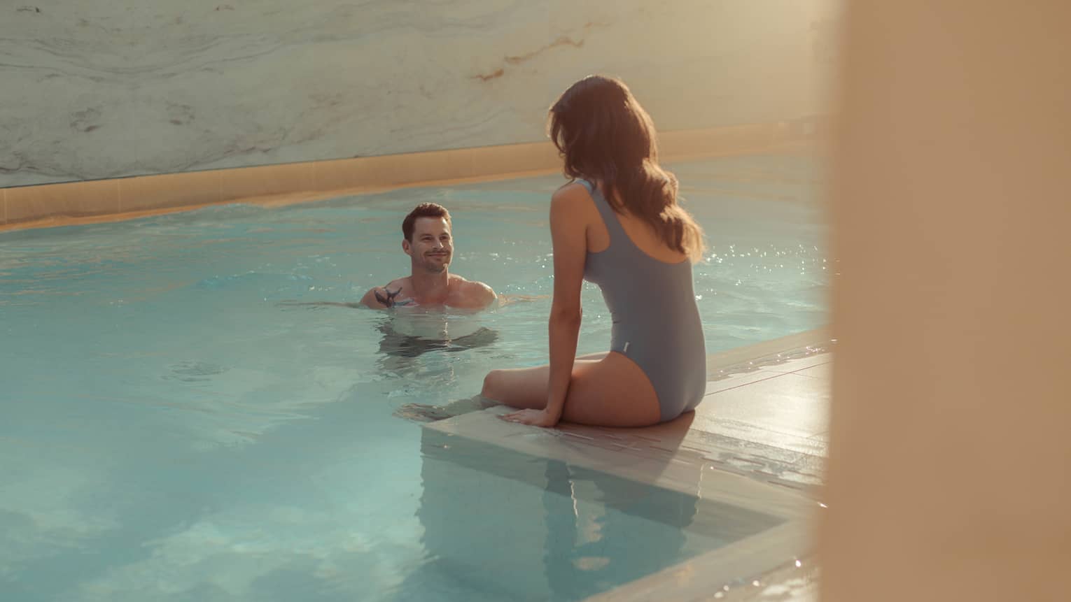 At an indoor pool with marble walls illuminated by natural sunlight, a woman sits on the pool's tiled edge as a man stands in the clear, blue water facing her
