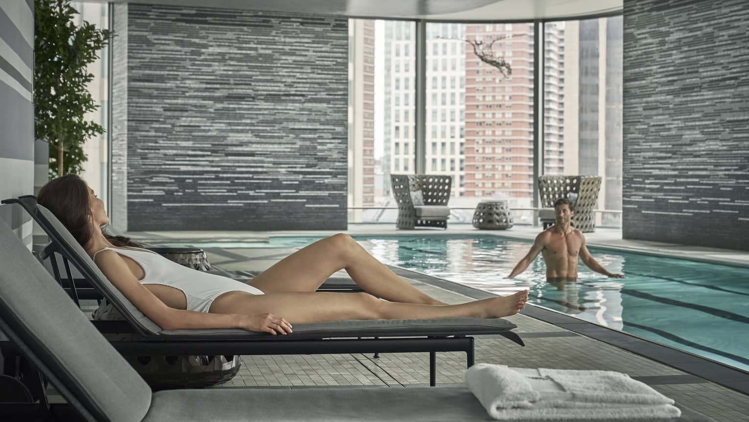 A woman in a white bathing suit and lounge chair looks on at her significant other in the lap pool