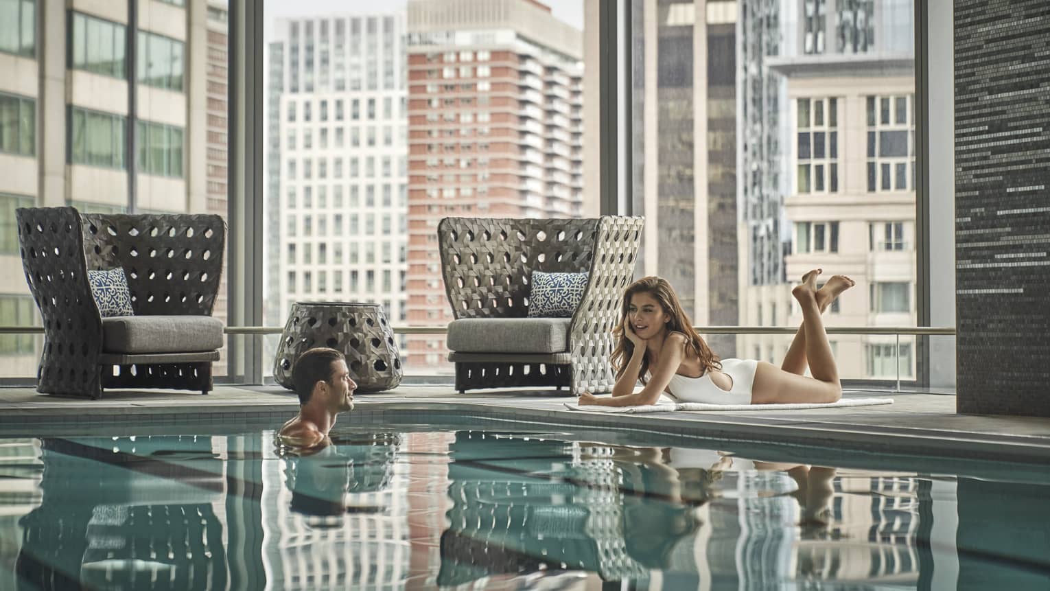 Woman in white bathing suit lounges poolside, talking to a man in the pool with the Boston skyline behind them 