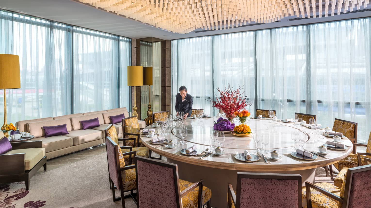 Server sets large round dining table by corner windows under ceiling covered with hanging lights