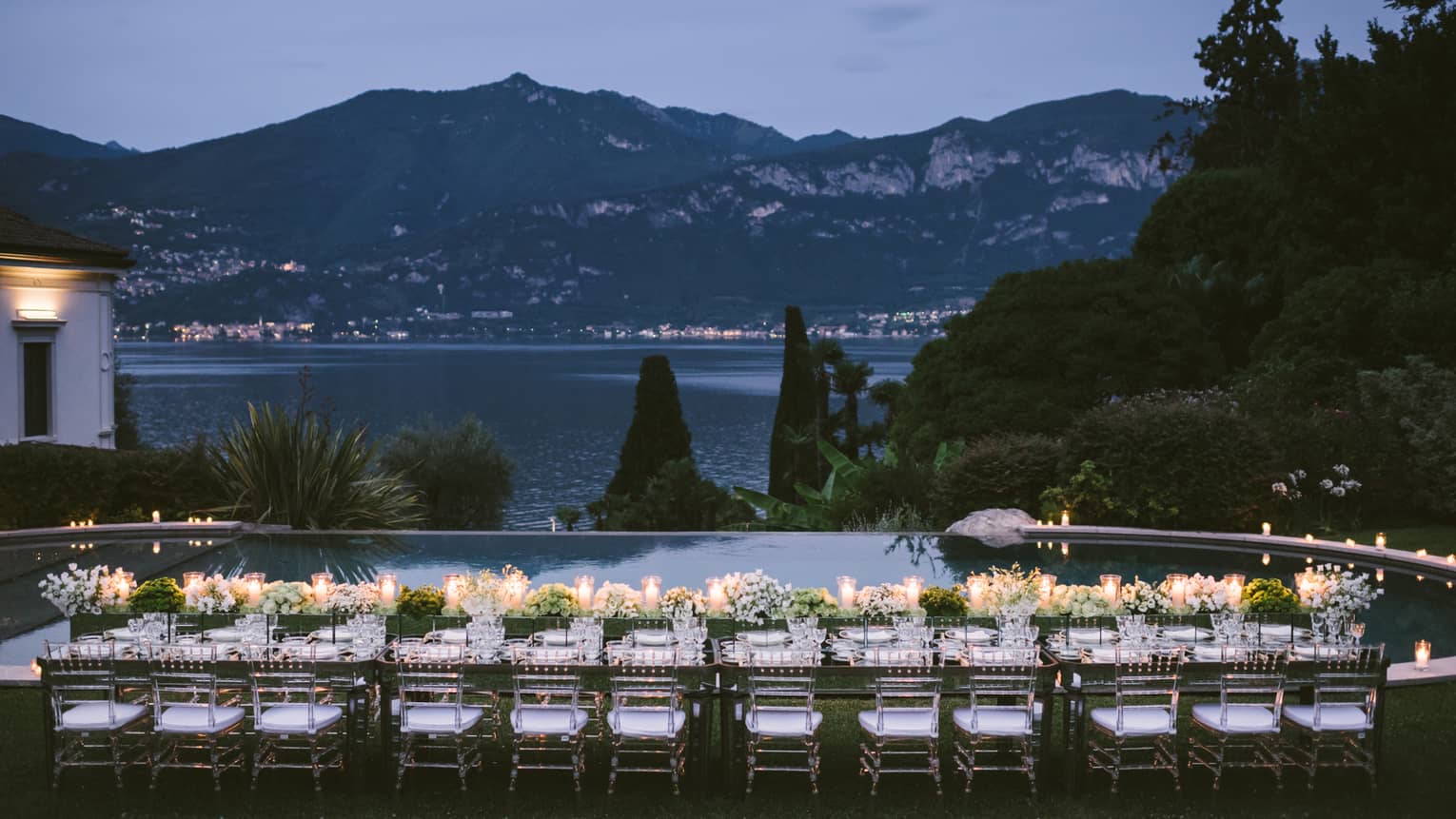 Long outdoor dining table with flowers, glowing candles by Lake Como, mountains at dusk