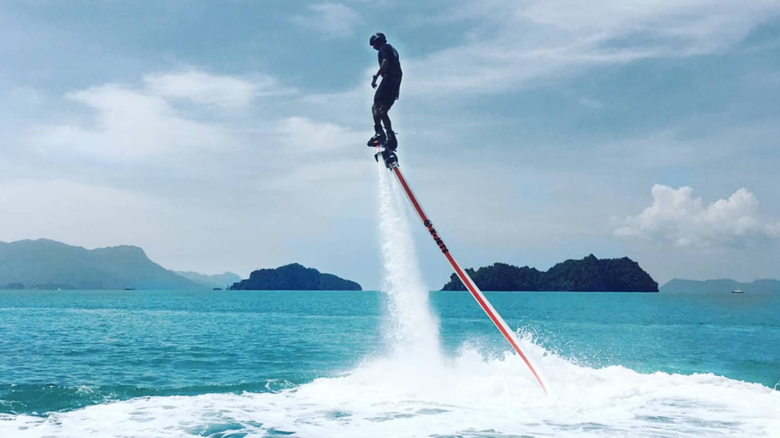 Water sprays as man soars above ocean attached to line, jet blade pack
