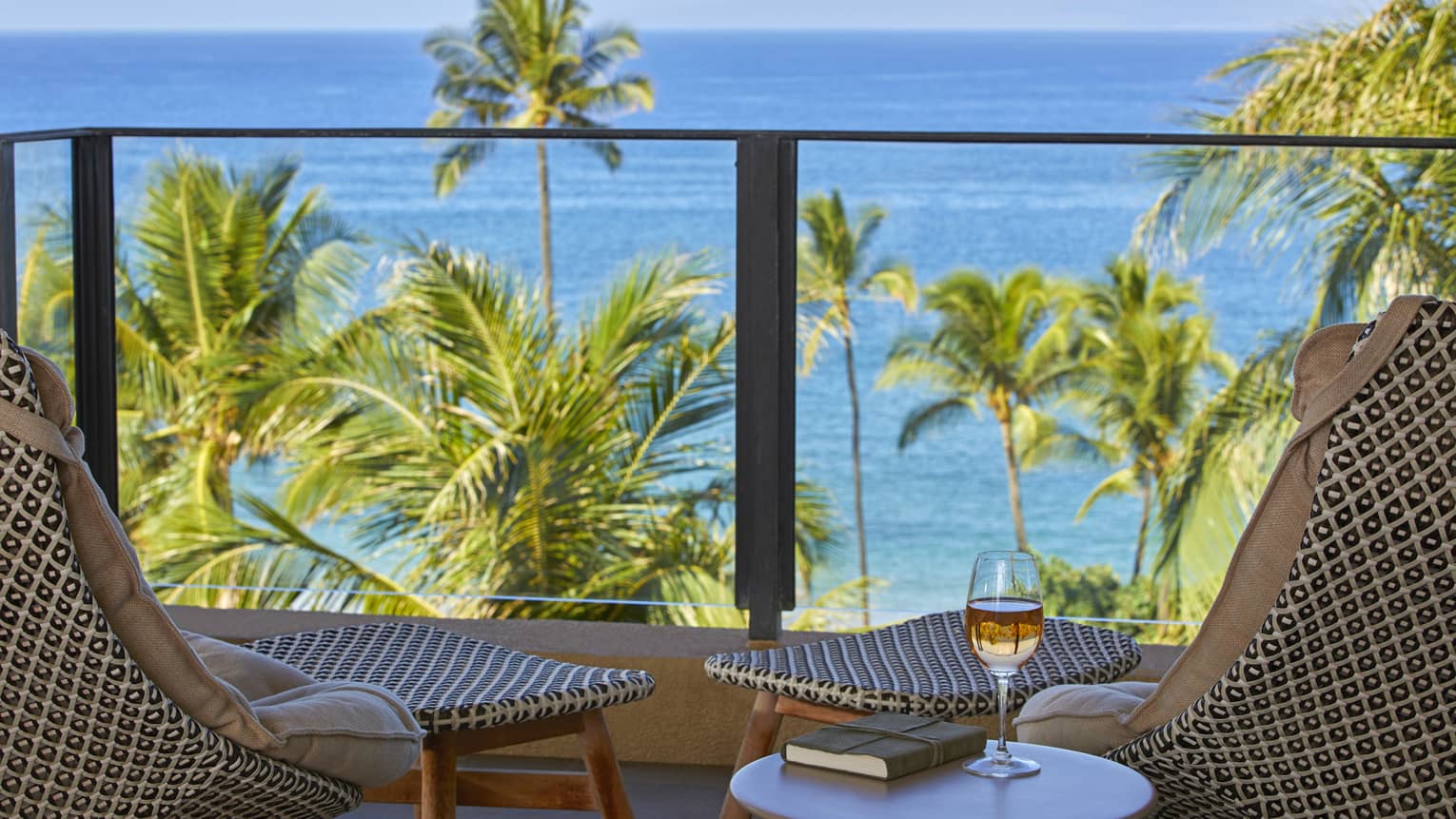 Elite suite balcony with two patterned chairs and a table with a glass of wine and book, overlooking palm trees and a tropical ocean view